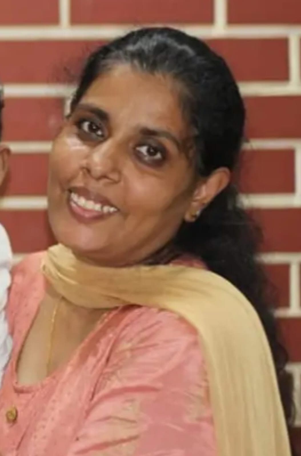 Smiling woman in a pink patterned kurta and yellow scarf posing against a brick wall.