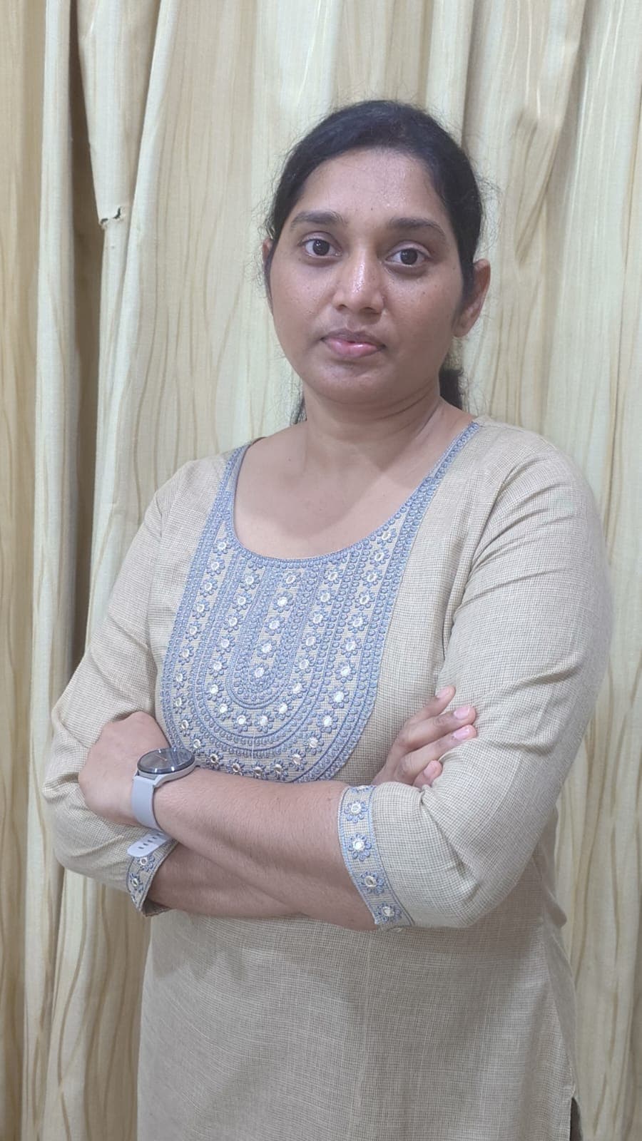 Woman with crossed arms wearing a beige embroidered kurta, standing against light-colored curtains.